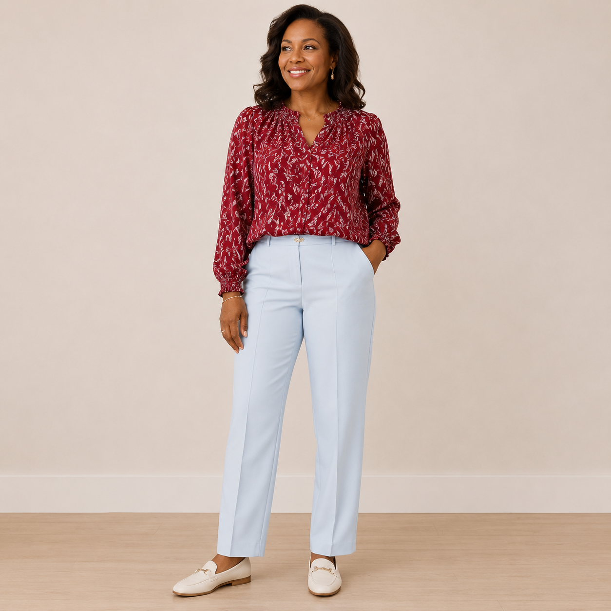 A woman stands in a studio wearing a long-sleeve red and white patterned blouse tucked into light blue straight-leg pants, with white loafers, against a beige wall and light wood floor.