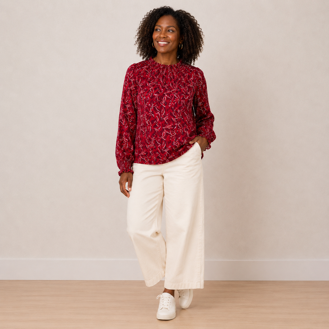 A woman modeling a long-sleeve red blouse with a white abstract pattern, paired with off-white wide-leg pants and white sneakers, standing against a neutral beige wall.