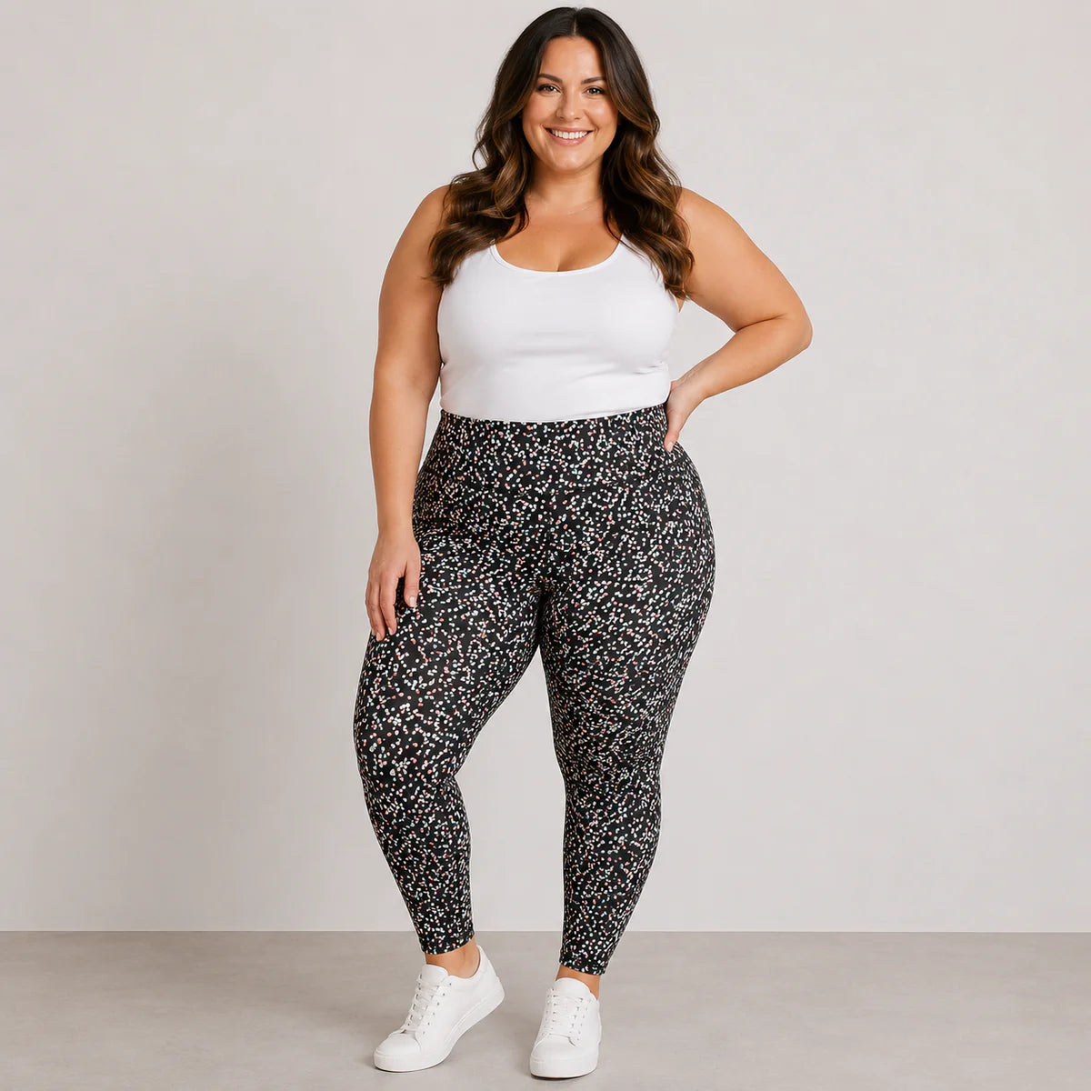 Woman wearing a white tank top and black patterned leggings on a plain background