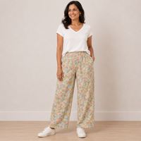A woman modeling a pair of wide-leg pants with a dense pastel floral print, paired with a white V-neck t-shirt and white sneakers, standing against a neutral beige wall.