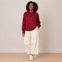 A woman modeling a long-sleeve red blouse with a white abstract pattern, paired with off-white wide-leg pants and white sneakers, standing against a neutral beige wall.