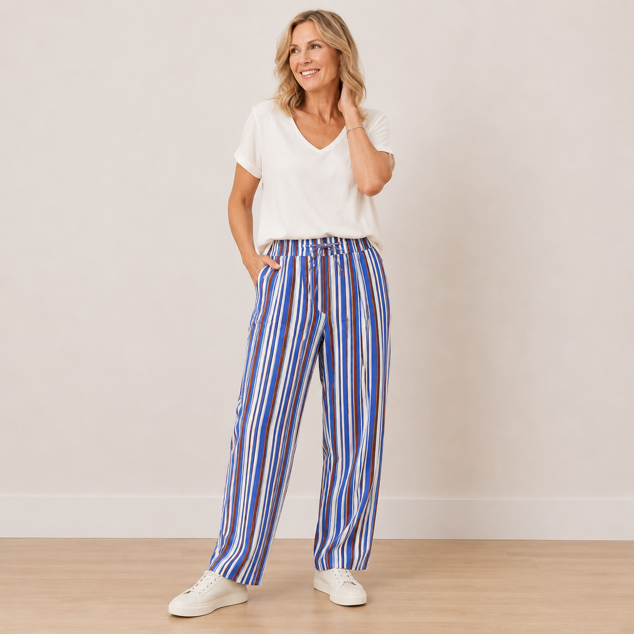 A woman modeling a pair of wide-leg trousers with vertical blue, red, and white stripes, paired with a white V-neck t-shirt and white sneakers, standing against a neutral beige wall.