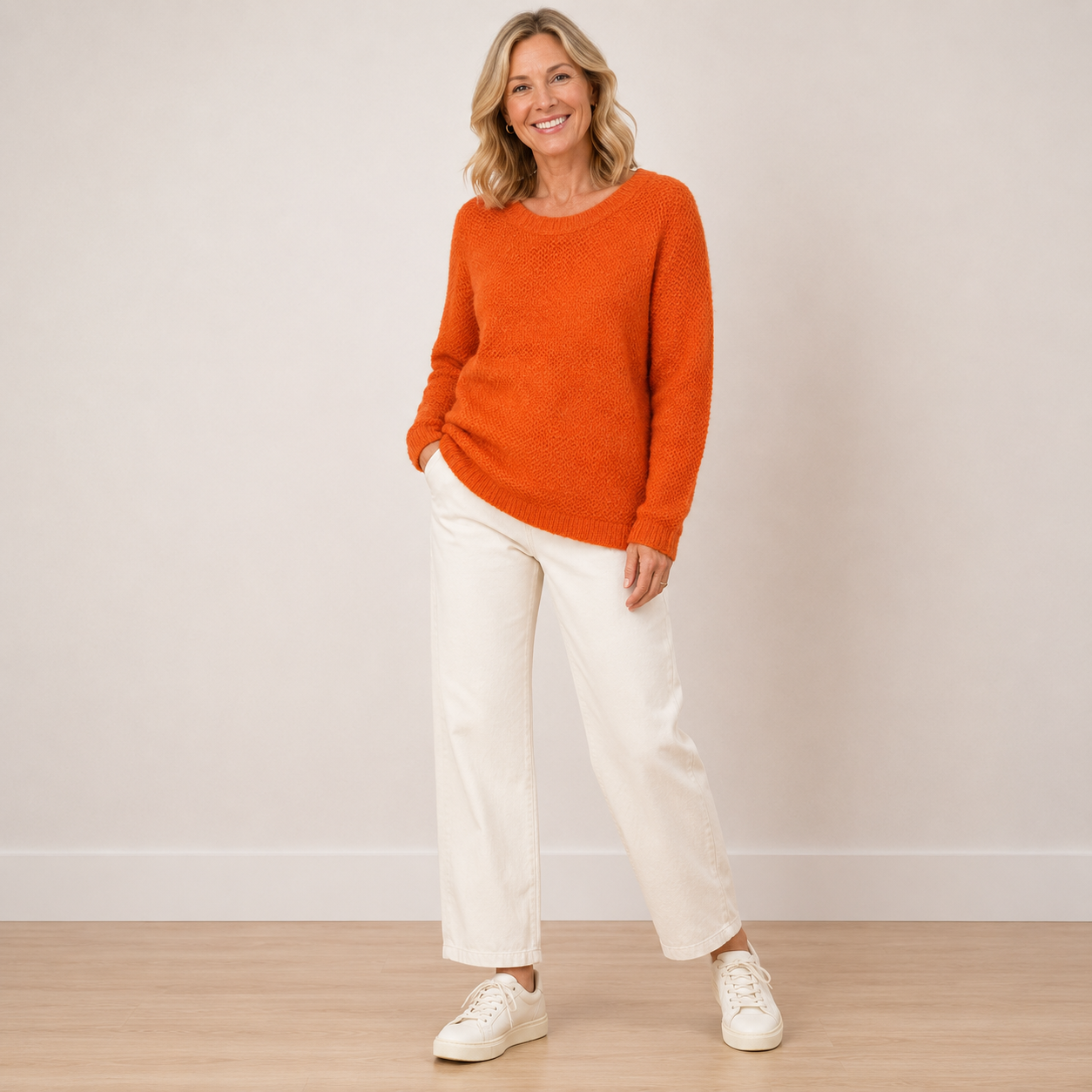 A woman stands in a studio wearing a fuzzy orange crewneck sweater, off-white wide-leg trousers, and white sneakers against a plain light beige wall and wood floor.
