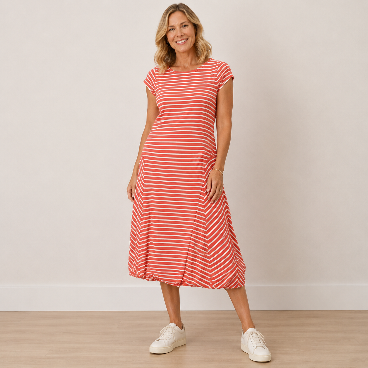 A woman modeling a red and white horizontally striped short-sleeve midi dress with a relaxed fit, paired with white sneakers, standing against a neutral beige wall and light wood floor.