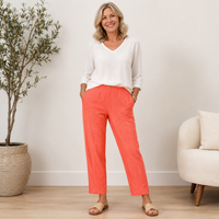A woman stands in a bright indoor room wearing a white V-neck long-sleeve top and coral-orange cropped pants with side pockets, paired with tan slide sandals.