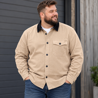 A man wearing a beige button-up overshirt with a black corduroy collar, styled with blue jeans over a white t-shirt, standing in front of a dark gray wooden wall.