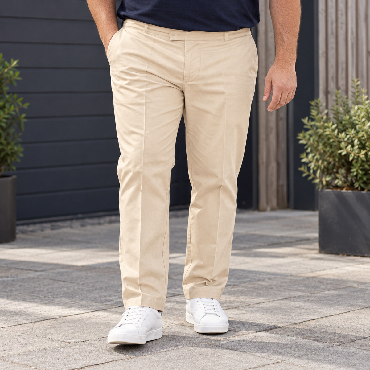 A man wearing beige chinos and white sneakers standing on a paved outdoor walkway in front of a dark gray wall.