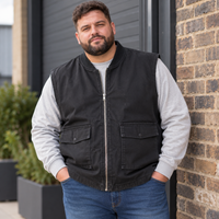 A man wearing a black zip-up sleeveless vest over a light gray long-sleeve shirt and blue jeans, standing outdoors against a brick wall and dark metal door.