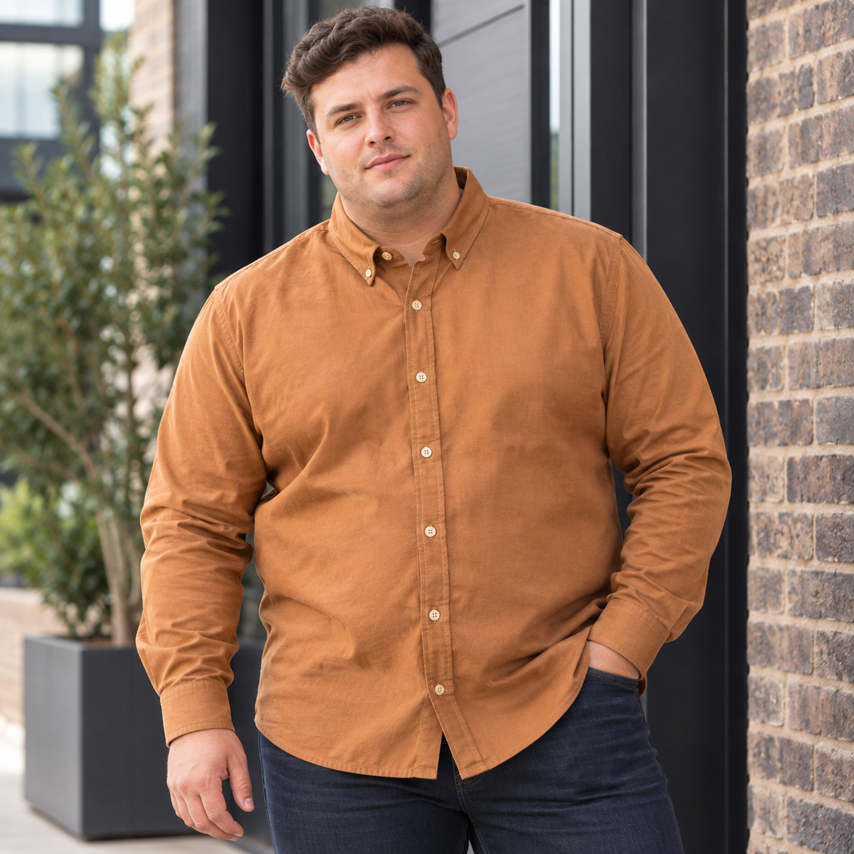 A man wearing a long-sleeve button-down shirt in a warm camel brown color, paired with dark jeans, standing outdoors in front of a brick wall and modern building entrance.