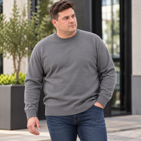 A man wearing a solid gray long-sleeve crewneck sweater and blue jeans, standing outdoors in front of a modern building with glass windows and potted plants.