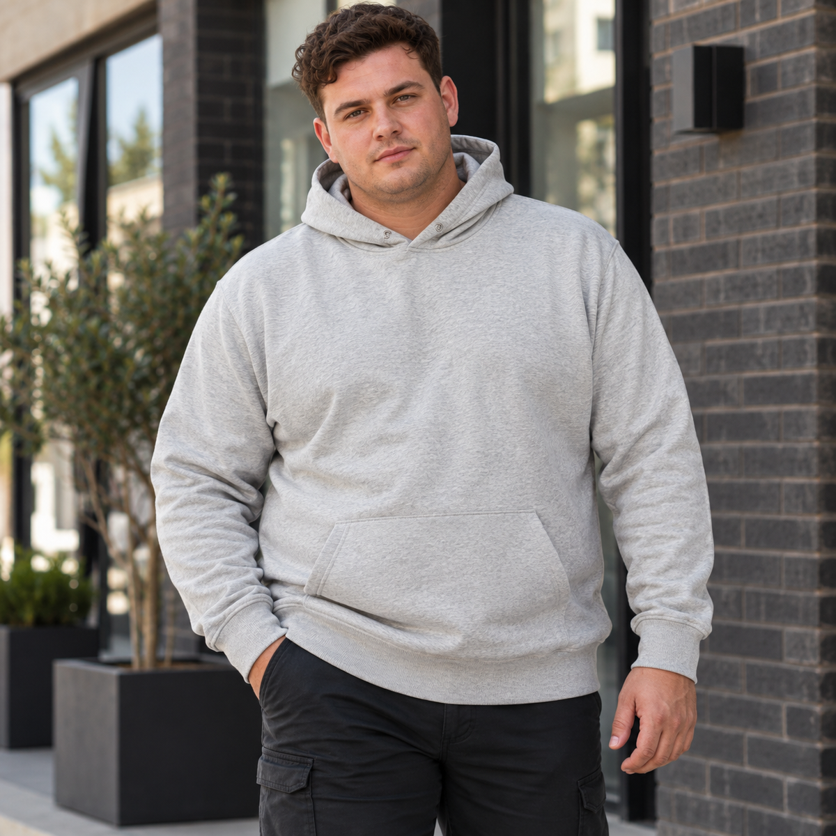 A man wearing a light gray heather pullover hoodie and dark cargo pants, standing outdoors in front of a modern brick building.