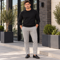 A man wearing a black crewneck sweater and light gray trousers stands outdoors on a paved walkway in front of a modern building with large windows and planters.