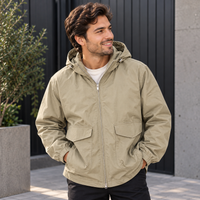 A man wearing a light khaki hooded zip-up jacket with front flap pockets, standing outdoors in front of a dark paneled wall.