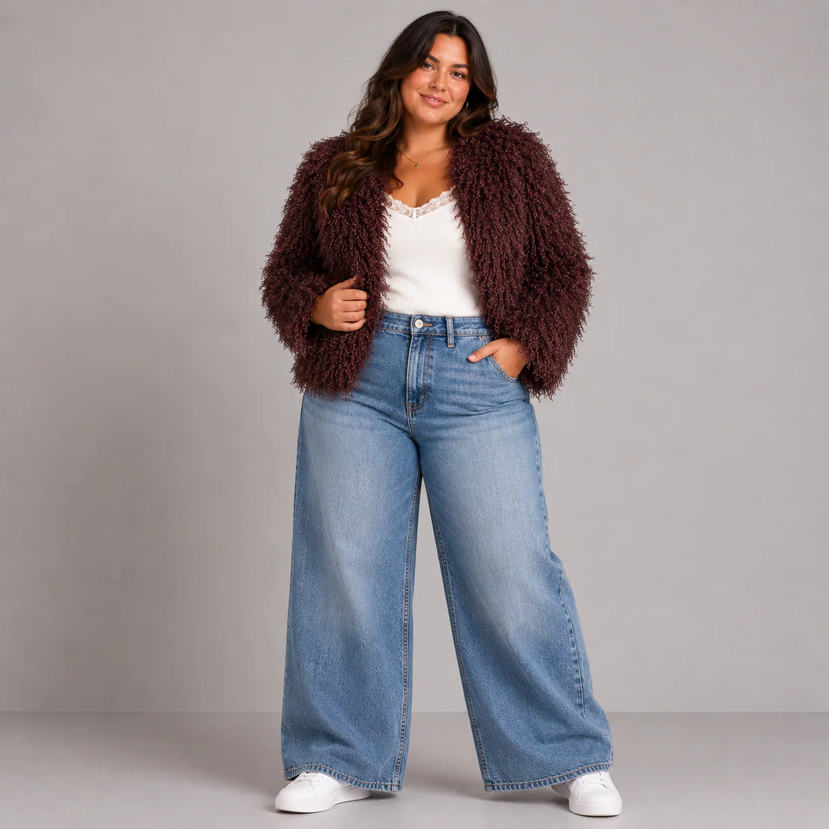 Woman wearing a brown fuzzy jacket, white top, blue jeans, and white sneakers on a gray background