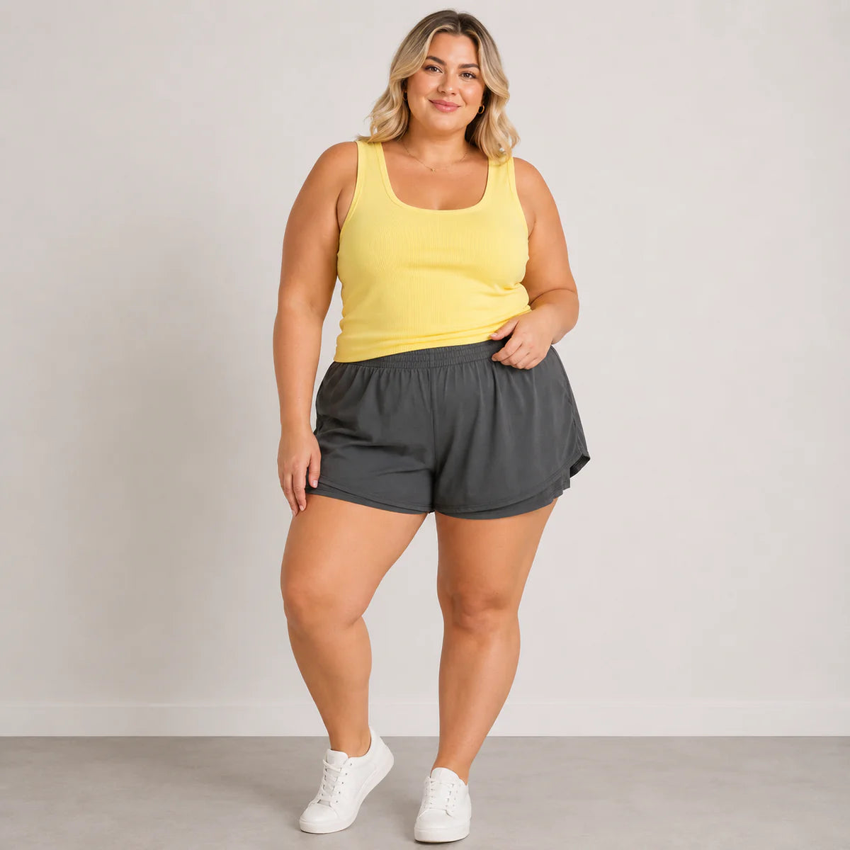 A plus-size woman modeling a yellow tank top and dark gray athletic shorts against a neutral studio background.