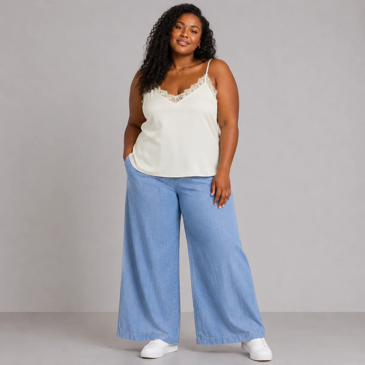 Woman wearing a white lace top and blue wide-leg pants against a gray background