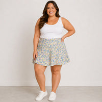A woman modeling a white sleeveless tank top and a short, flared floral skirt with white sneakers, standing against a plain light gray studio background.