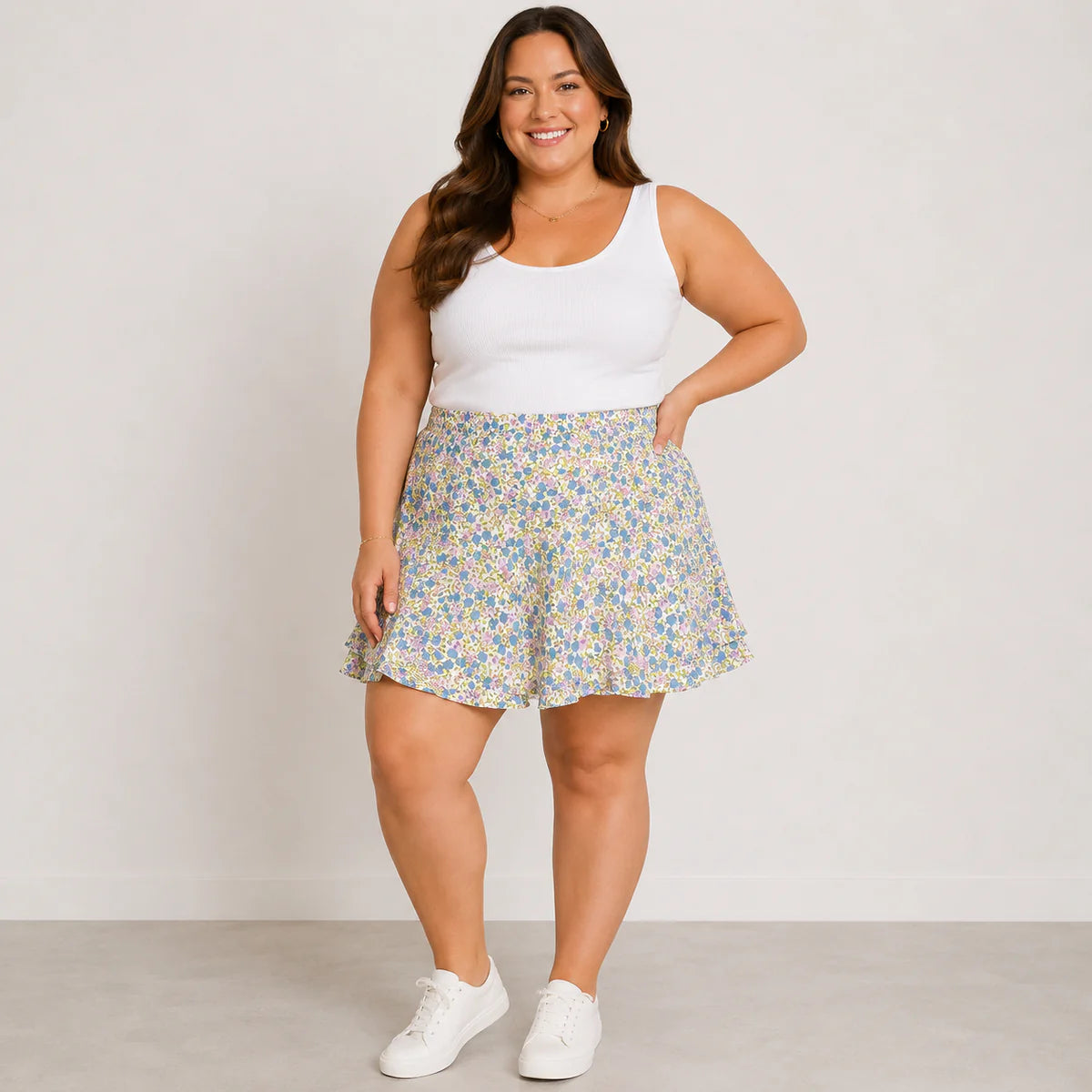 A woman modeling a white sleeveless tank top and a short, flared floral skirt with white sneakers, standing against a plain light gray studio background.
