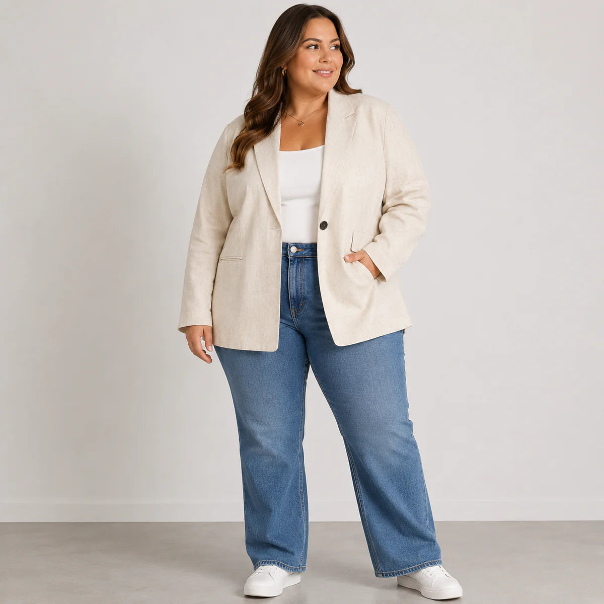 A woman modeling a light beige blazer over a white top and blue wide-leg jeans, standing against a plain light gray studio background.