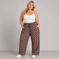 A woman modeling wide-leg brown pants with a white polka dot pattern, paired with a white tank top and white sneakers, against a solid gray studio background.